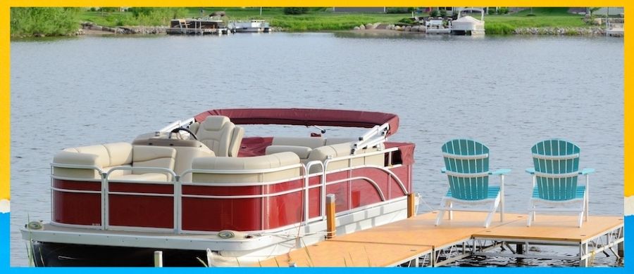 A red and tan pontoon boat sits at the dock on a lake. It is on small lake, with a small dock. 2 chairs sit on the end of the dock.