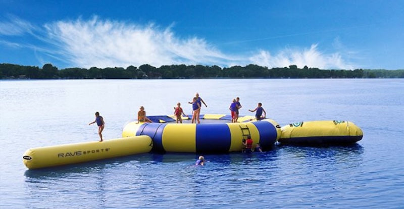 Best Water Trampoline and Best Water Bouncer Buyer's Guide. Image shows a blue and yellow inflatable water trampoline on the middle of a lake. There are 5 people playing on the water trampoline on a beautiful day with only a few clouds in the sky.