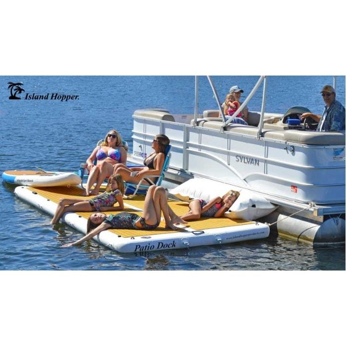 Side corner view of the Island Hopper Patio Dock Floating Swim Platform from a slight distance. The inflatable floating dock is attached to a pontoon and there are several people suntanning and sitting in chairs on the inflatable dock. There are also 2 people in the pontoon and the water is dark blue.